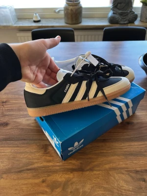 Adidas Samba  - Klassiska Adidas Samba sneakers i svart och vitt med ikoniska tre ränder på sidan och gummisula. Ovandelen är i mocka och läder, med vita detaljer och svart bas. Perfekta för dig som gillar retrostil och streetwear. Skön passform och tidlös design. Helt nya. Fel storlek därför säljer jag. Hör av er vid minsta fundering. Först till kvarn.