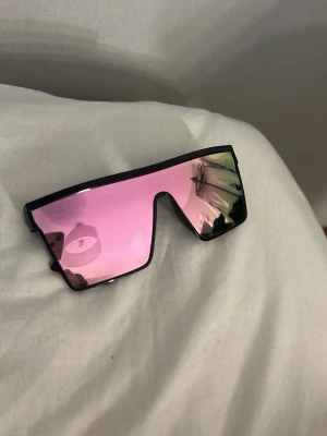 Svarta solglasögon med rosa spegelglas - Säljer ett par trendiga solglasögon med svart båge och stora, rektangulära rosa spegelglas. Glasögonen har en futuristisk vibe och passar perfekt för dig som vill sticka ut med din stil.