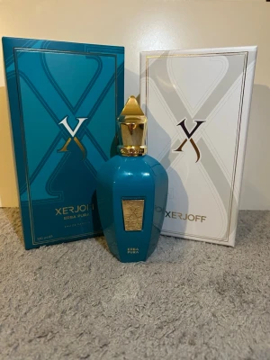 Xerjoff Erba Pura Eau de Parfum 100ml - Lyxig parfym från Xerjoff, Erba Pura, i en elegant turkos flaska med gulddetaljer och exklusiv förpackning. Flaskan har en unik form och pryds av en guldfärgad etikett och lock. Kommer i en snygg box med färgstark insida och stilren design.