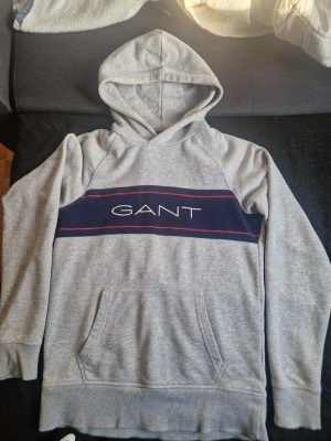 GANT Hoodie - Snygg grå hoodie från GANT med stor blå panel och vit logga över bröstet, samt röda detaljer. Klassisk känguruficka framtill och mjukt material. Perfekt för en chill och stilren look. (Har ett pytte litet hål men syns knapp)