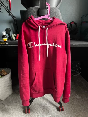Champion hoodie - Snygg röd hoodie från Champion med stor vit logga på bröstet och vita dragsnören i huvan. Klassisk känguruficka framtill och ribbade muddar vid ärmar och nederkant. Perfekt för en chill och sportig look.