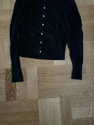 Svart kofta med knappar - Stilren svart kofta med långa ärmar och guldiga knappar framtill. Klassisk rak passform och mjukt material. 