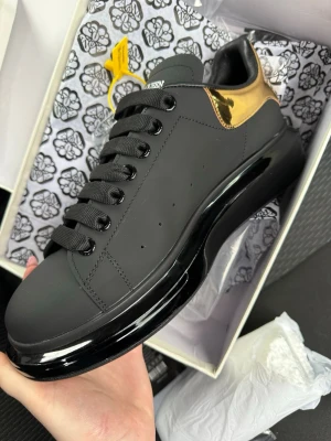 Svarta sneakers Alexander McQueen - Stilrena svarta sneakers från Alexander McQueen med chunky svart sula och metallic guld-detalj på hälen. Skorna har breda svarta skosnören och är tillverkade i slätt läder. Perfekta för dig som vill ha en statement sneaker med lyxig känsla.