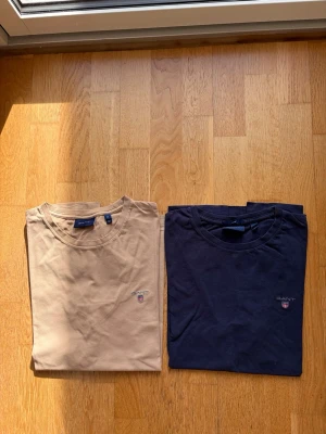 GANT t-shirts i beige och marinblå - Säljer två stilrena t-shirts från GANT, en i beige och en i marinblå. Båda har rund hals, korta ärmar och den klassiska GANT-loggan broderad på bröstet. Perfekta basplagg i mjuk bomull som funkar till allt och särskilt på sommaren med ett par snygga shorts! 🎉Beigea T-shirten har ett litet hål på ryggen därav är den såld för endast 20 kr!(se sista bild) 🎉Marinblå T-shirten har lite urtvättad färg men fortfarande som ny därav 80 kr för den! 🎉För mer info kom dm!