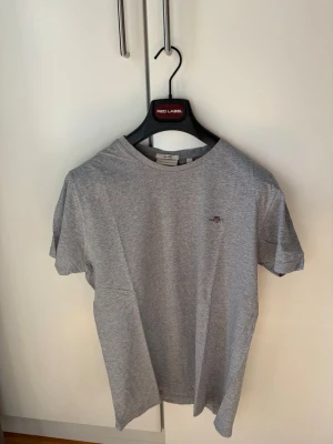 GANT: T-Shirt - Snygg grå t-shirt från GANT i storlek M. Klassisk modell med rund hals och liten broderad logga på bröstet. Tillverkad i mjuk bomull som känns skön mot huden. Perfekt till jeans eller shorts för en clean och enkel stil. 🎉Slim Fit  🎉För mer info skriv dm!