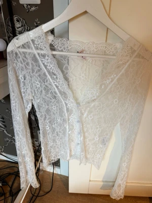 Vit spetsblus med v-ringning - Säljer en superfin vit blus i spets med v-ringning och långa ärmar. Blusen har ett romantiskt blommigt spetsmönster och är genomskinlig, perfekt att styla med ett linne under. Snygg detalj med vågig kant längs ringningen.