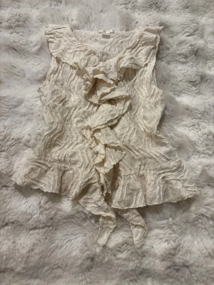 Krämvit volangblus med knyt - Supersöt krämvit blus med volanger framtill och knytband nedtill. Blusen är ärmlös och har ett vågigt, strukturerat tyg som ger en cool och unik look. Perfekt att styla med jeans eller kjol för en trendig outfit.