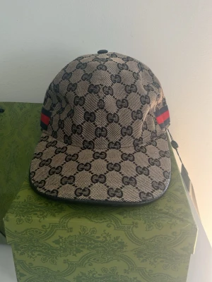 Gucci keps med GG-mönster - Säljer en beige keps från Gucci med klassiskt GG-mönster i svart och detaljer med röd och blå rand på sidan. Kepsen har böjd skärm och är tillverkad i canvasmaterial. Perfekt för dig som vill ha en lyxig och trendig accessoar.
