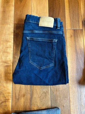 Mörkblå jeans från GANT - Säljer ett par stilrena GANT jeans  Märke: GANT  Färg: Mörkblå  Passform: Slim  Storlek: 170cm  GANT är ett premiumvarumärke känt för sin tidlösa, klassiska stil med rötter i amerikansk "preppy"-mode. Deras jeans är designade för att hålla länge och passa i alla sammanhang – både till vardag och mer uppklädda outfits.  Skick: Bra använt skick – inga hål eller större skador, endast normalt slitage.  Perfekta för dig som vill ha ett par snygga märkesjeans som funkar till allt.  Hör av dig vid intr