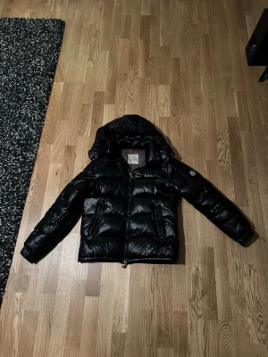 Svart dunjacka från Moncler - Säljer en svart dunjacka från Moncler med glansig finish och avtagbar huva. Jackan har dragkedja framtill, två sidofickor och klassisk Moncler-logga på ärmen. Insidan är grå med detaljer och etiketter. Perfekt för kalla vinterdagar.