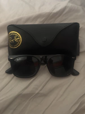 Svarta Ray-Ban Wayfarer solglasögon - Klassiska svarta Ray-Ban Wayfarer solglasögon med mörka glas och ikonisk logga på linsen. Snygg och tidlös design som passar till allt. Kommer med original svart fodral med Ray-Ban-märke.