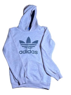 Grå hoodie från Adidas med logga - Snygg grå hoodie från Adidas med stor svart Trefoil-logga på bröstet. Klassisk känguruficka framtill och ribbade muddar vid ärmar och midja. Tillverkad i mjukt bomullsmaterial och har en rymlig huva. Perfekt för en avslappnad streetwear-look.