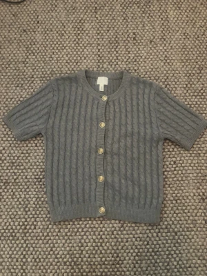 Grå kabelstickad kofta med kort ärm - Snygg grå kabelstickad kofta med korta ärmar och rund hals. Koftan har ribbade muddar och fem guldfärgade knappar framtill. Perfekt att slänga över en topp för en chill look.