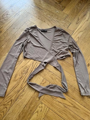 Bolero - Säljer en beige croppad bolero  i ribbad polyester. Toppen har långärmad design, v-ringning och knytband som ger en snygg passform. Perfekt för dig som gillar trendiga och bekväma plagg.