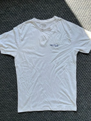 Handbroderad vit t-shirt från Denim Project - Vit t-shirt från Denim Project. På bröstet finns en handbroderad text 'MATT 6:33' i blått. ”Men sök först Guds rike och hans rättfärdighet, så skall ni få allt det andra också.” Det finns gult smuts på framsidan. 