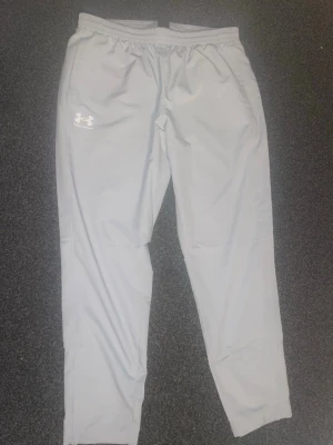 Ljusgrå träningsbyxor Under Armour - Snygga ljusgrå träningsbyxor från Under Armour med elastisk midja och logga på vänster lår. Perfekta för gymmet eller löprundan. Tillverkade i ett lätt och bekvämt material som andas.