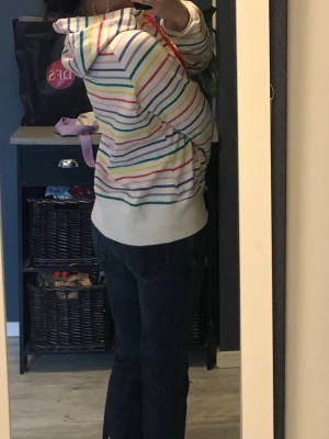 Vit hoodie med färgglada ränder - Säljer en vit hoodie med dragkedja och färgglada ränder i blått, rött, gult och grönt. Hoodien har röda snören i huvan och två fickor framtill. Perfekt för dig som gillar en lekfull och färgstark stil.