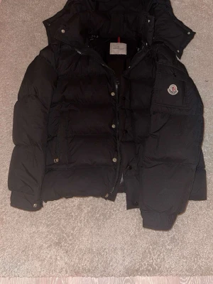Svart dunjacka från Moncler - Säljer en svart dunjacka från Moncler med klassisk puffer-design och huva. Jackan har dragkedja och knappar framtill, samt en ficka med Moncler-logga på ärmen. Insidan har ett serietidningsmönster och glansigt foder. Perfekt för kalla vinterdagar.