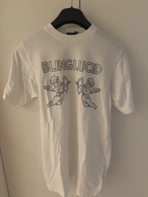 Vit BLINGLUCID t-shirt med tryck - Vit t-shirt från BLINGLUCID med coolt tryck av två amor-figurer och texten BLINGLUCID framtill. Klassisk rund hals och korta ärmar. Perfekt för dig som gillar streetwear och vill sticka ut med ett unikt motiv.