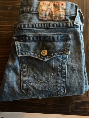 Blå True Religion jeans med ficklock - Säljer ett par klassiska blå jeans från True Religion med raka ben och ikoniska ficklock bak. Jeansen har kontrastsömmar och en patch med logga bak i midjan. Perfekta för dig som gillar streetwear och vill ha en unik detalj på dina jeans. Skriv om ni har några frågor💕