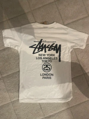 Vit Stüssy t-shirt med tryck - Säljer en vit t-shirt från Stüssy med ikoniskt svart logotryck på bröstet och stort tryck på ryggen med städer som New York, Los Angeles, Tokyo, London och Paris. Klassisk passform och rund hals. Perfekt för dig som gillar streetwear och enkel stil.