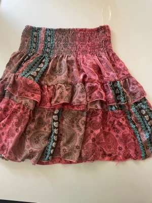 Rosa volangkjol med paisleymönster - Söt kortkjol i rosa toner med paisley- och blommönster samt turkosa detaljer. Kjolen har bred smock i midjan och flera volanger för en bohemisk look. Tillverkad i tunn bomull som känns lätt och luftig. Kjolen är köpt utomlands så märke vet jag ej, skriv privat för fler bilder🌴💓