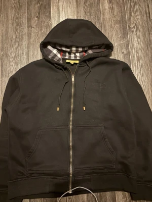 Svart hoodie från Burberry med dragkedja - Säljer en svart hoodie från Burberry med dragkedja och klassiskt rutigt foder i huvan. Hoodien har två fickor framtill, justerbara snören i huvan och diskret Burberry-logga broderad på bröstet. Perfekt för dig som gillar stilrena plagg med exklusiva detaljer.