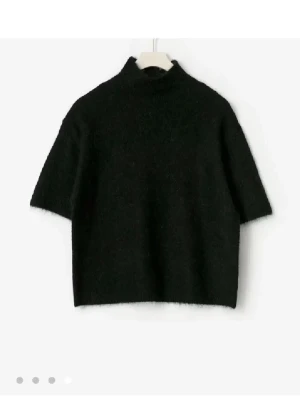 Svart stickad tröja med halv polo - En svart stickad tröja med halv polokrage och korta ärmar. Tröjan har en boxig passform och är gjord i ett mjukt, fluffigt material som ger en trendig look. Perfekt att styla lager på lager eller bära som den är.