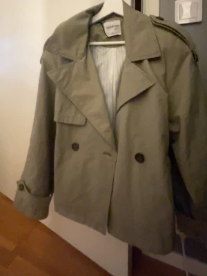 Grön trenchcoat  - Stilren kort trenchcoat i grön från Bershka med dubbelknäppning, bred krage och axelklaffar. Jackan har knytskärp i midjan och dekorativa knappar vid ärmsluten. Tillverkad i ett lätt bomullsliknande material, perfekt för lager på lager.