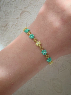 Handgjort blommigt pärlarmband – Pastell & Guld – Daisy Chain - Handgjort pärlarmband med små blommor (daisies). Perfekt för vårens och sommarens outfits eller som en fin present! • Design: Alternerande blommor i mintgrönt/turkost och ljusgult med vita mittpunkter. Mellan varje blomma sitter guldfärgade pärlor • Detaljer: Guldfärgat klolås med förlängningskedja för justerbar passform. • Stil: Boho, somrigt, cottagecore och feminint. • Skick: Helt nytt/oanvänt. Passar både till vardags och fest. Armbandet är lätt och bekvämt at