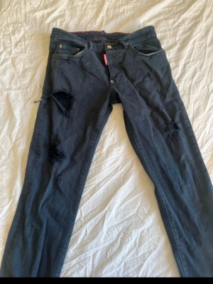 Svarta  jeans från Dsquared2 - Säljer ett par svarta jeans från Dsquared2 med flera hål på benen för en edgy look. Jeansen har klassisk femficksmodell, knappgylf och röda detaljer vid linningen. Materialet är denim med tydlig struktur och passformen är rak.