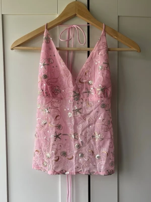 Rosa paljettbroderad linnetopp - Supersöt rosa linnetopp med tunna axelband och knyt i nacken. Toppen har v-ringning och är dekorerad med glittriga paljetter och broderade blommor i silver och guld. Toppen har en öppen rygg med knytning. Perfekt för dig som vill sticka ut lite extra!