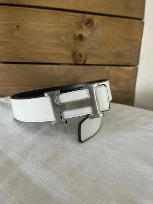 Vändbart svart och vitt skärp med H-spänne - Snyggt vändbart skärp i svart och vitt med ett stort H-format metallspänne. Skärpet har en stilren design och är tillverkat i skinn, vilket ger en exklusiv känsla. Perfekt accessoar för att lyfta din outfit. 115cm