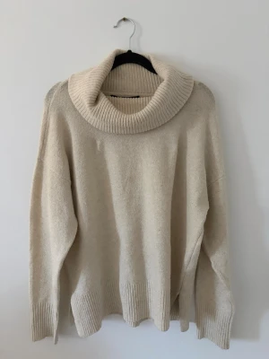 Beige polotröja från Vero Moda - Mysig beige stickad polotröja från Vero Moda med bred, ribbad krage och ribbade muddar vid ärmslut och nederkant. Oversized passform som ger en avslappnad look. Perfekt för kalla dagar och enkel att matcha med jeans eller kjol.