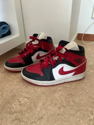 Nike Air Jordan 1 Mid röd/svart/vit - Säljer ett par Nike Air Jordan 1 Mid sneakers i klassisk röd, svart och vit färgkombination. Skorna har läderöverdel, röda snören och den ikoniska swoosh-loggan på sidan. Perfekta för dig som gillar streetwear och vill sticka ut.
