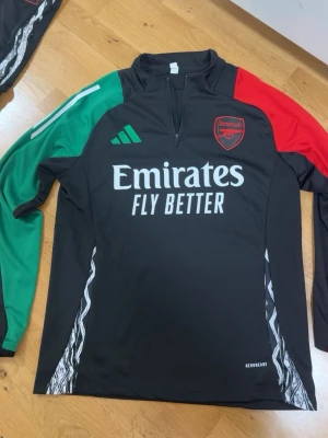 Arsenal träningströja Adidas svart - Snygg svart långärmad träningströja från Adidas med Arsenal-logga på bröstet. Tröjan har grönt och rött på ärmarna, vit Adidas-logga och Emirates Fly Better-tryck framtill. Tillverkad i funktionsmaterialet Aeroready, perfekt för träning.