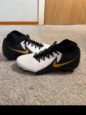 Nike fotbollsskor och benskydd - Säljer fotbollsskor från Nike, endast använda en gång. De är vita och svarta med guld detaljer. Storlek 42. Nypriset är 1400