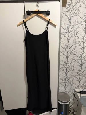 Svart långklänning med smala axelband - En stilren svart långklänning med smala axelband och rak passform. Klänningen har en enkel och minimalistisk design som gör den lätt att styla. Perfekt för dig som gillar en clean look.