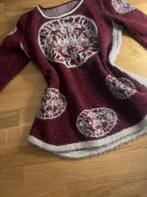 Vinröd stickad tröja med mönster - Mysig vinröd stickad tröja med vita, runda mönster och kontrasterande vita kanter. Tröjan har rund halsringning och långa ärmar. Materialet ser fluffigt och mjukt ut, perfekt för kalla dagar.