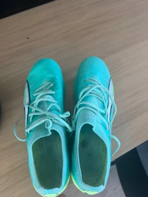 Turkosa Puma fotbollsskor - Säljer ett par turkosa Puma fotbollsskor med vita och svarta detaljer samt gula inslag vid hälen. Skorna har snörning och dobbar för spel på gräs. Ovandelen är i syntetmaterial som ger bra bollkontroll och lätt känsla på foten. Perfekta för dig som vill sticka ut på planen!