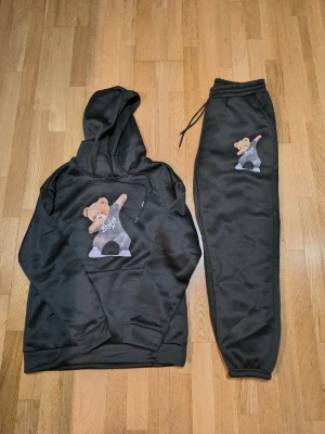 Svart set med björnmotiv - Säljer ett svart set med luvtröja och byxor, båda med tryck av en söt björn som dabbar. Setet har en avslappnad passform och är gjort i mjukt material, perfekt för en chill look. Dragsko i midjan på byxorna och fickor på tröjan.