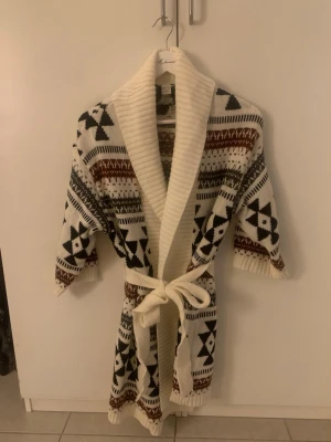 Mönstrad kofta från H&M Divided - Mysig morgonrock från H&M Divided i storlek XS. Vit bas med svart och brunt geometriskt mönster, bred ribbad krage och knytskärp i midjan. Perfekt för att chilla hemma i. Stickad känsla och trekvartsärmar. Oanvänd och i väldigt bra skick, oversize modell