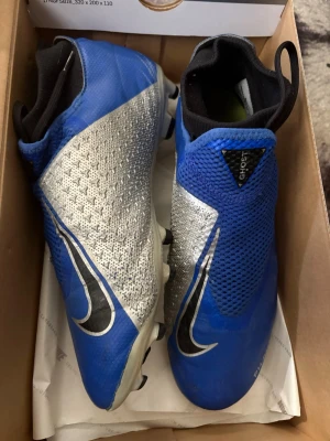 Nike Phantom Vision blå fotbollsskor - Säljer ett par Nike Phantom Vision fotbollsskor i blått och silver med svarta detaljer. Skorna har en strumpliknande och en stor Nike Swoosh på sidan. Ovandelen är i syntetmaterial med struktur för bättre bollkontroll. Perfekta för dig som vill sticka ut på planen. Skorna passar även 42,5