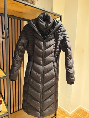 Svart lång dunjacka med huva - Säljer en superfin svart lång dunjacka från Guess med huva och dragkedja framtill. Jackan har quiltad design, markerad midja och ett litet Guess-emblem på ärmen. Sparsamt använd. Storlek S. 