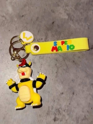 Super Mario Bowser nyckelring - Gul och svart nyckelring med Bowser-figur från Super Mario. Nyckelringen har ett gult silikonband med färgglad Super Mario-text och en rund vit bricka med gul detalj. Perfekt accessoar för Mario-fans!