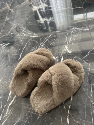 Beiga fluffiga korsade tofflor - Supersköna tofflor i beige fluffigt material med korsad design över foten. Mjuka och gosiga, perfekta för att chilla hemma. Sula och ovandel är helt täckta av det mjuka materialet för extra komfort.
