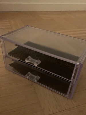 sminkförvaringslåda med lådor - Praktisk och stilren sminkförvaringslåda i transparent plast med två utdragbara lådor. Lådorna har svarta halkskyddsmattor som håller dina sminkverktyg och tillbehör på plats. Perfekt för att organisera borstar, pennor och småprylar på sminkbordet.