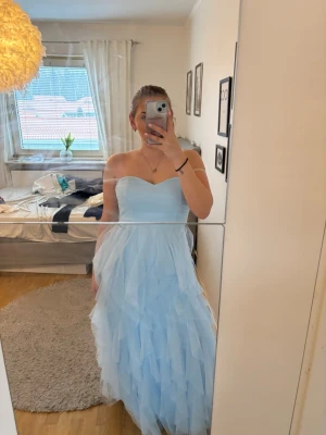 Ljusblå axelbandslös långklänning - Säljer en ljusblå axelbandslös långklänning med vacker volang och flera lager av tunn tyll. Klänningen har en hjärtformad urringning och är perfekt för speciella tillfällen. Endast använd 1 gång.