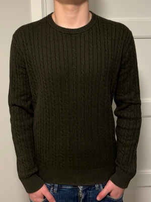 Mörkgrön kabelstickad tröja från Riley - Snygg mörkgrön kabelstickad tröja från Riley med rund halsringning och långa ärmar. Tröjan har ett klassiskt flätmönster och ribbade muddar vid ärmslut och nederkant. Perfekt för dig som gillar stilrena och tidlösa plagg. Hör av dig vid minsta intresse!✨⭐️💫
