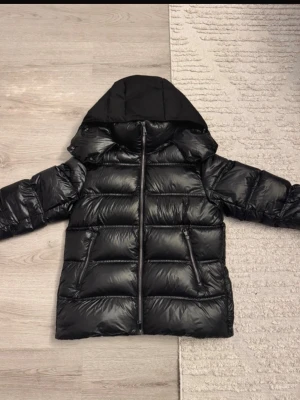 Svart pufferjacka med huva - Snygg svart pufferjacka med glansig finish och stor huva. Jackan har två dragkedjeförsedda fickor framtill och breda, vadderade paneler som ger en riktigt puffig look. Perfekt för kalla vinterdagar och riktigt trendig stil.